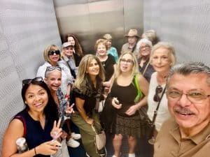 Smiling group of adults in a narrow elevator posing for a selfie toward the camera, all looking cheerful.