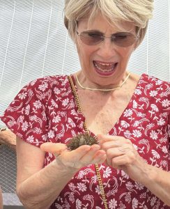 Older woman in a red floral dress holds a spiky sea urchin in cupped hands, wearing sunglasses and a gold necklace, at an outdoor display.