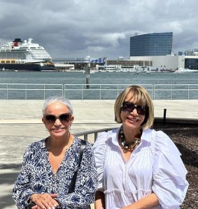 Two smiling women pose at a waterfront with a large cruise ship and city buildings in the background.