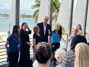 A man in a suit speaks to a small group of women on a waterfront balcony with palm trees in the background.