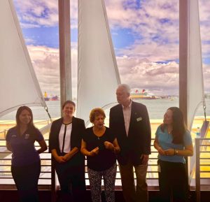 Five people stand in a line on a ship deck with tall white sails, overlooking the water and a distant cruise ferry in the background.