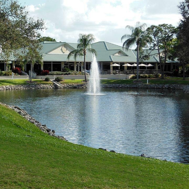 Pond with a central fountain in a landscaped park; a light-colored building with palm trees in the background.